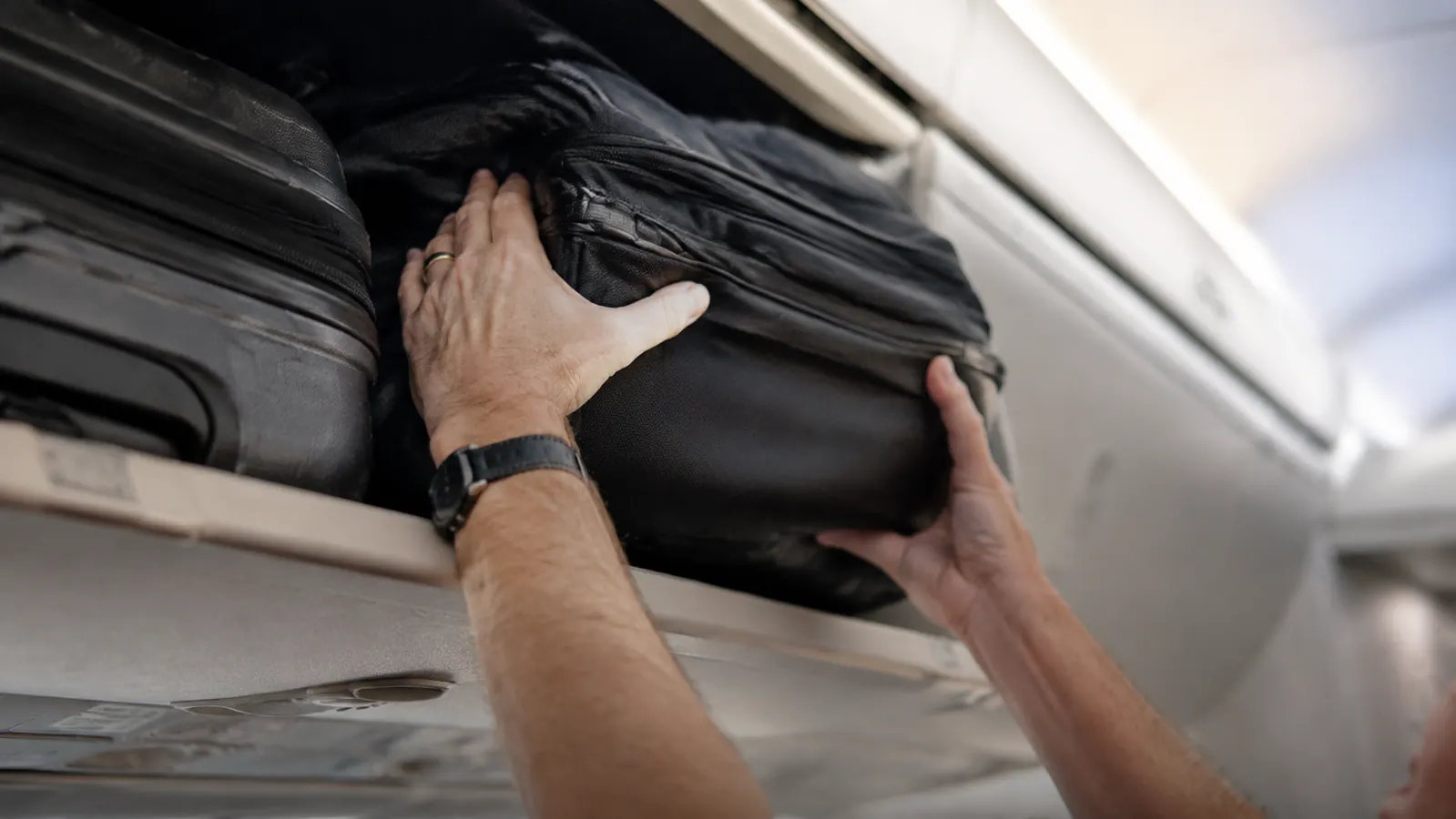 Traveler wrestling a carry-on bag into an overhead bin on an airplane, bag visibly overstuffed