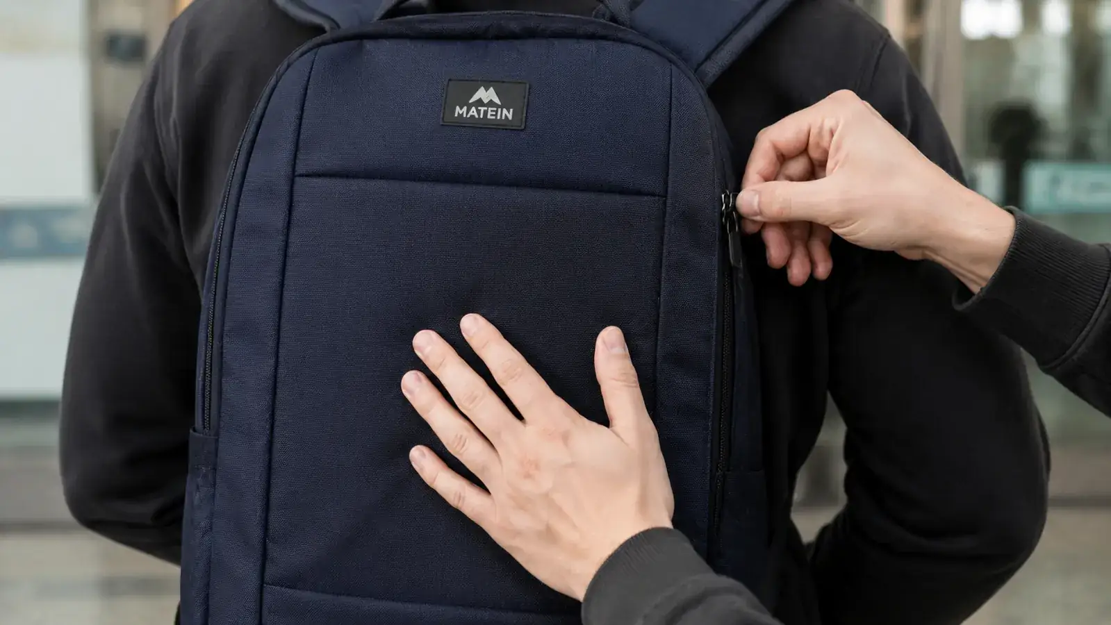 Close-up of a person's hand testing the back-panel hidden zipper on a dark backpack while another hand tries to reach it from behind