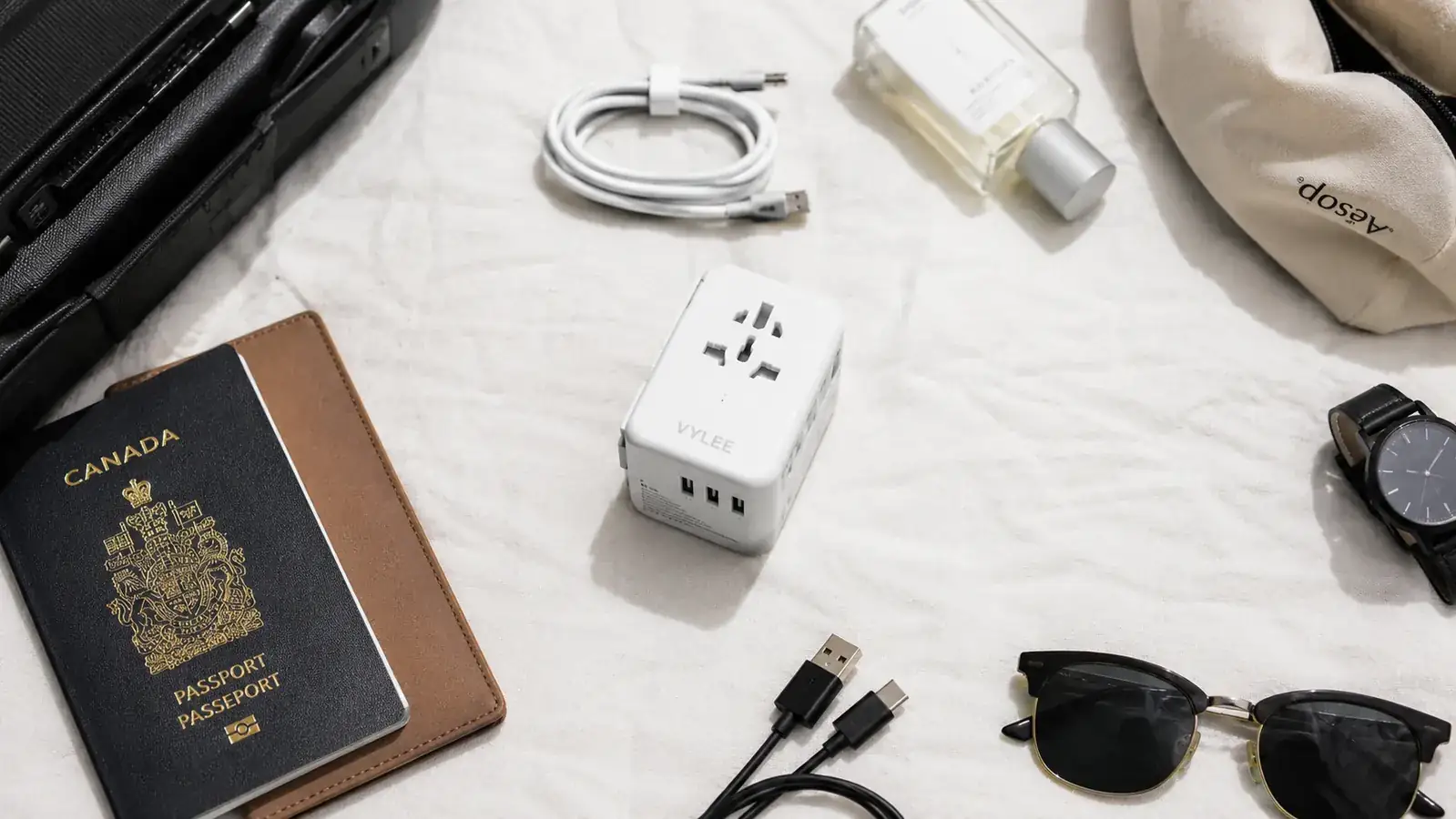 Flat lay of a minimalist travel packing setup showing the VYLEE adapter among passport, cables, and a small carry-on bag